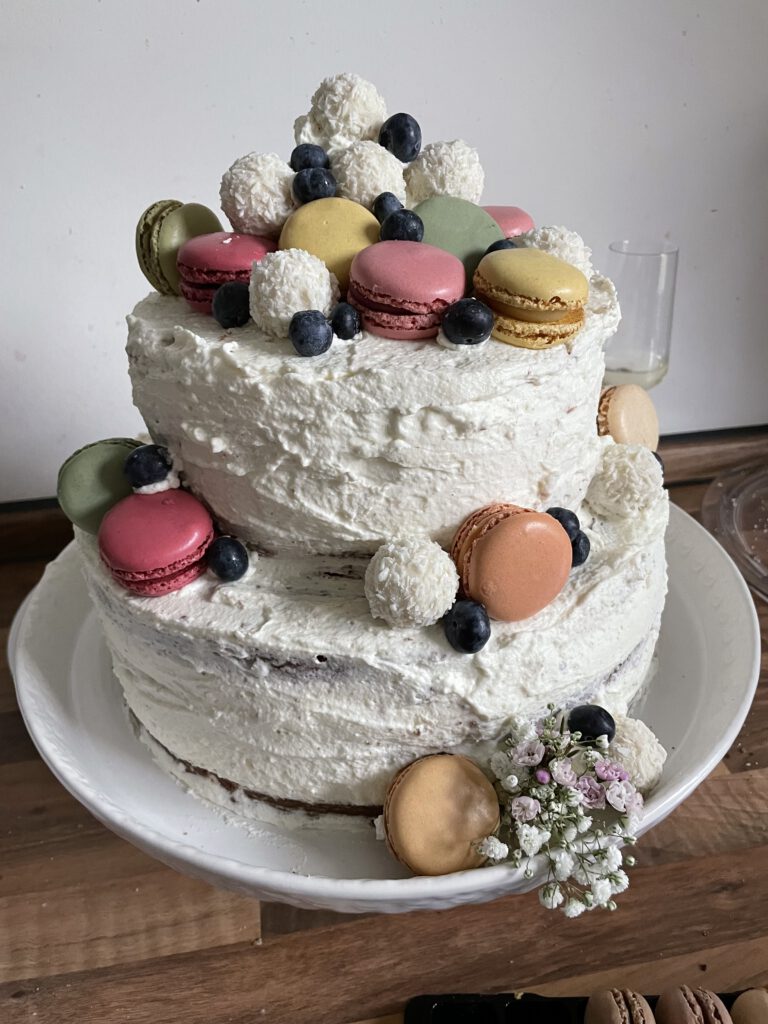 Eine zweistöckige Torte mit weißer Creme ummantelt und mit bunten Maccarons, Blaubeeren und Raffaello dekoriert.