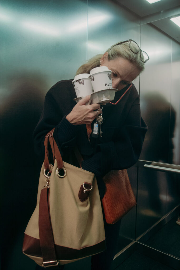 Eine Frau steht im Aufzug, hält Coffee to go becher, autoschlüssel und tasche in der hand, während sie telefoniert und in der Tasche etwas sucht.